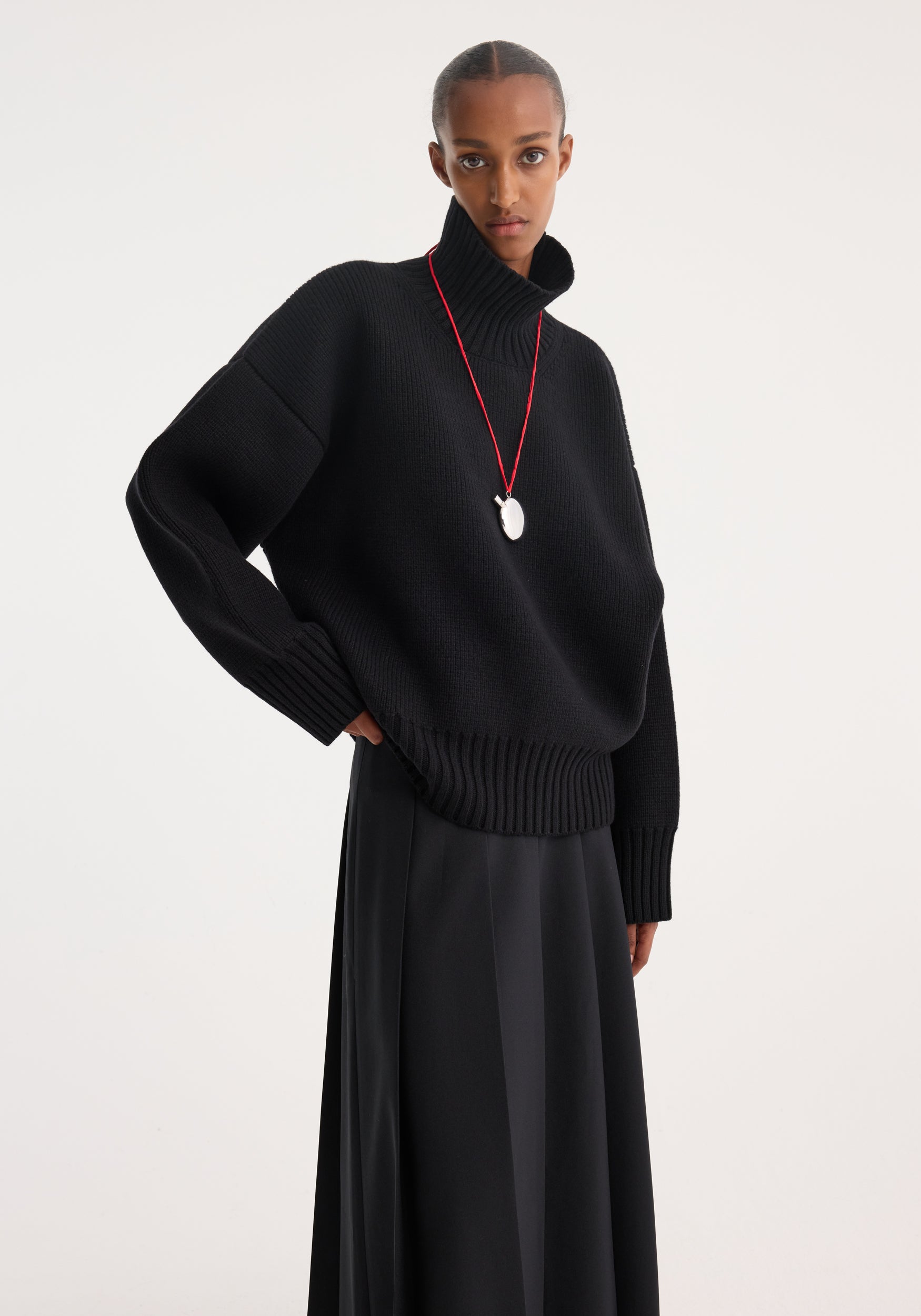 Model wearing a black Sculptural compact turtleneck | black sweater with a high ribbed collar and long sleeves, paired with a black skirt and a long necklace with a white pendant.