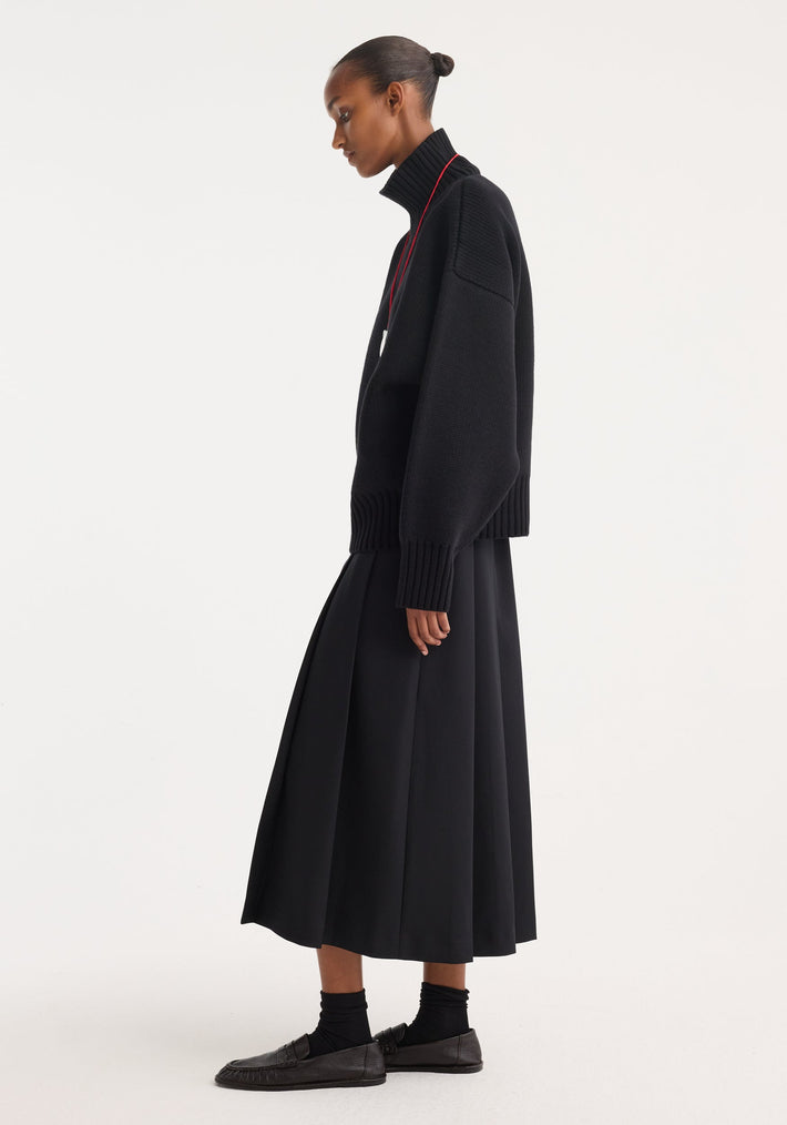 Model in profile wearing Sculptural compact turtleneck in black, showcasing its chunky knit, relaxed fit, dropped shoulders, paired with a pleated midi skirt.