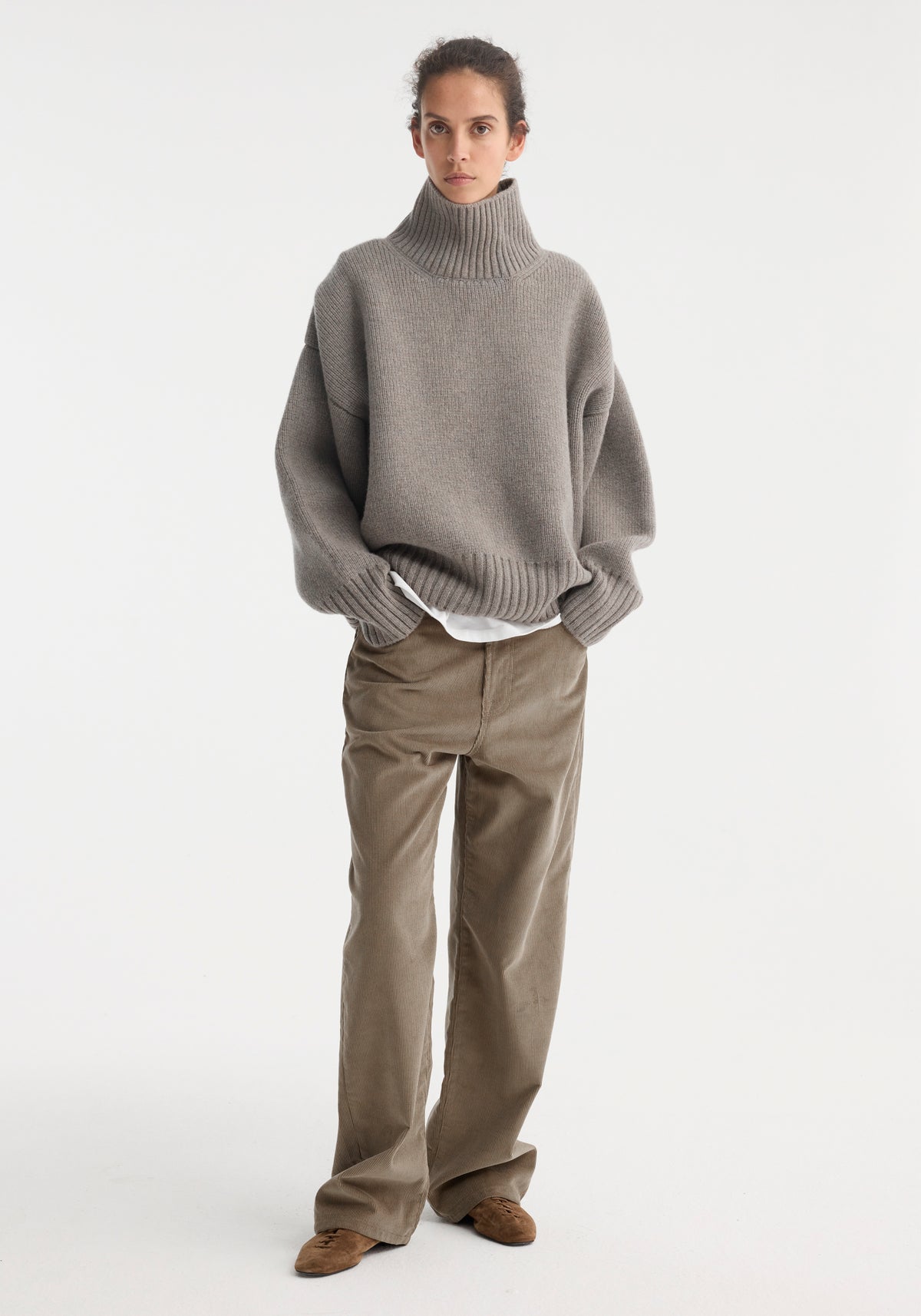 Woman wearing a loose-fitting Sculptural compact turtleneck | clay melange sweater with a high ribbed collar, paired with wide-leg brown pants and brown shoes, standing against a white background.