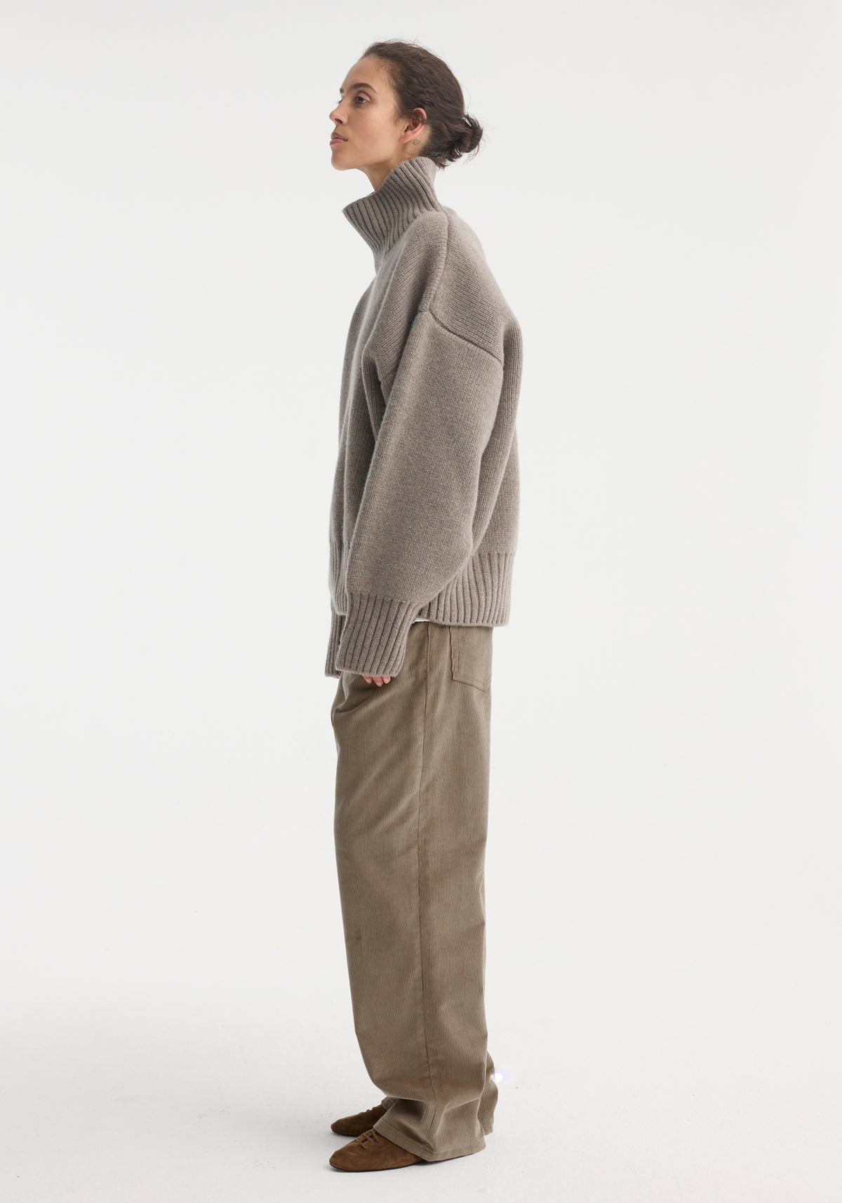 Profile view of a woman wearing a Sculptural compact turtleneck | clay melange with long sleeves and a high ribbed neckline, paired with loose-fitting earth-toned pants and brown shoes against a plain white background.