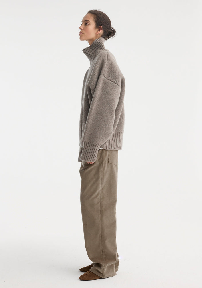 Woman wearing Sculptural compact turtleneck | clay melange, an oversized grey knit with drop shoulders, over loose taupe trousers, styled with brown suede lace-up shoes.