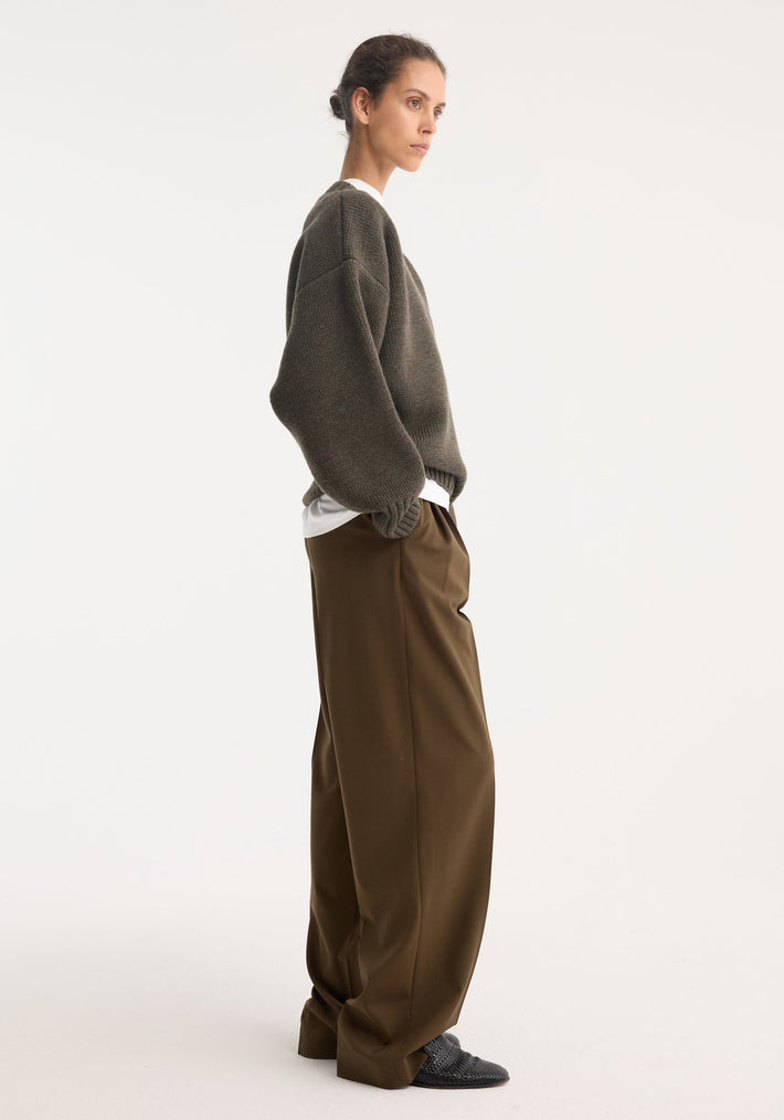 Person in profile wearing Sculptural compact v-neck sweater, oversized and chunky-knit, over a white shirt, paired with wide-leg trousers and slip-on shoes. Hair in low bun.