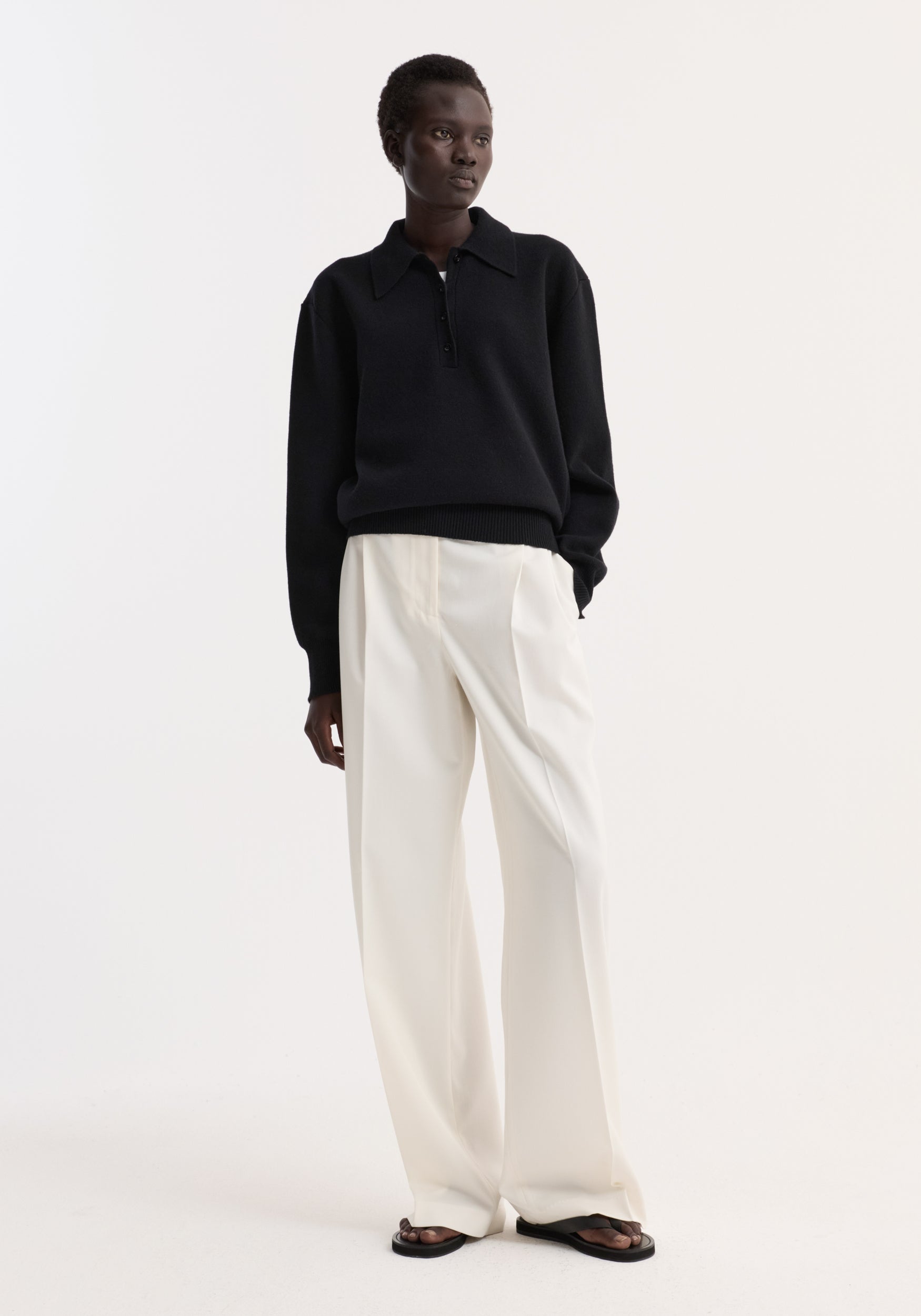Model wearing a black compact knitted polo | black with long sleeves and a collar, paired with white wide-leg trousers and black sandals, standing against a plain white background.