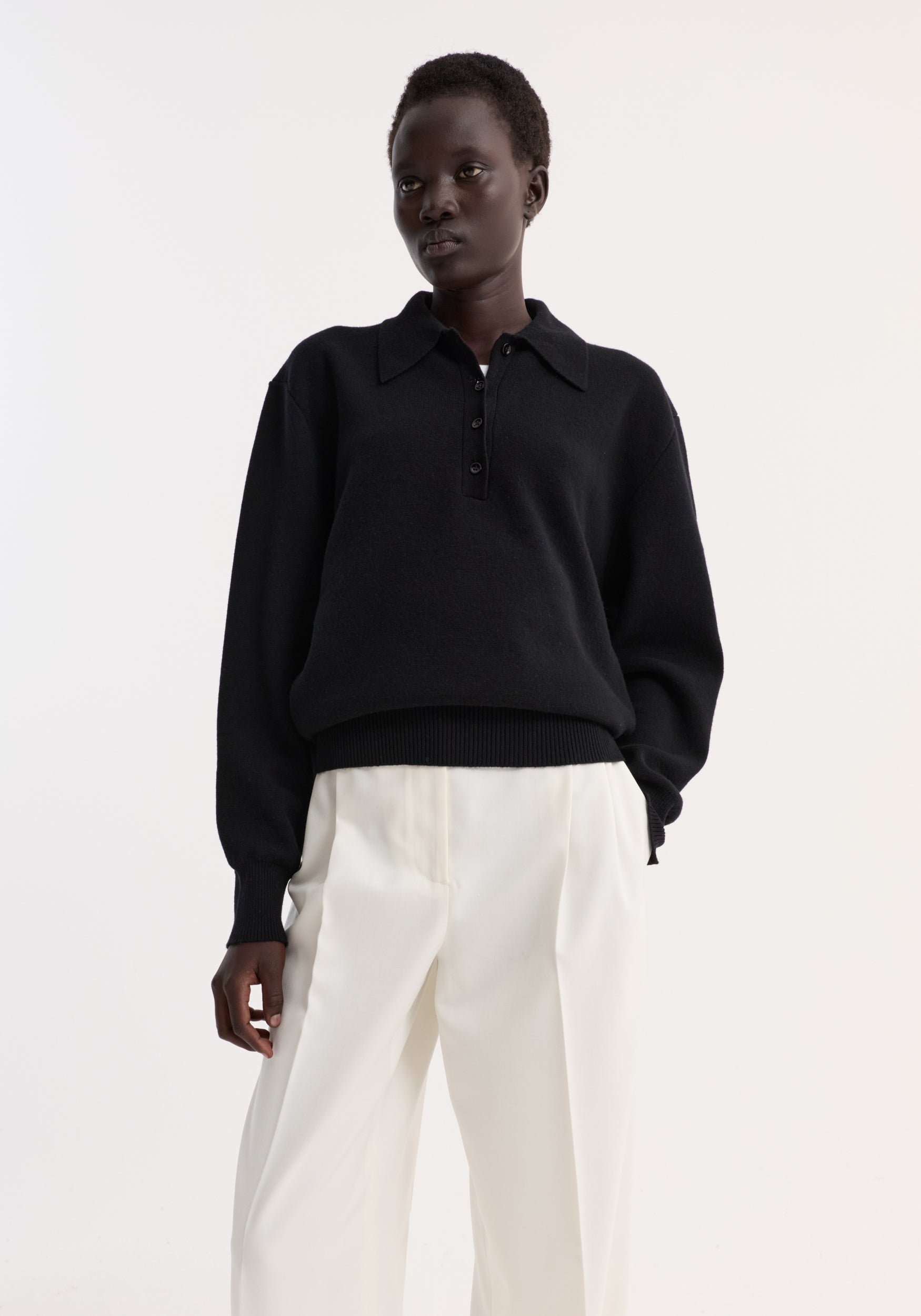 A model wearing a black Compact knitted polo | black with long sleeves and a buttoned collar paired with loose white trousers against a plain white background.