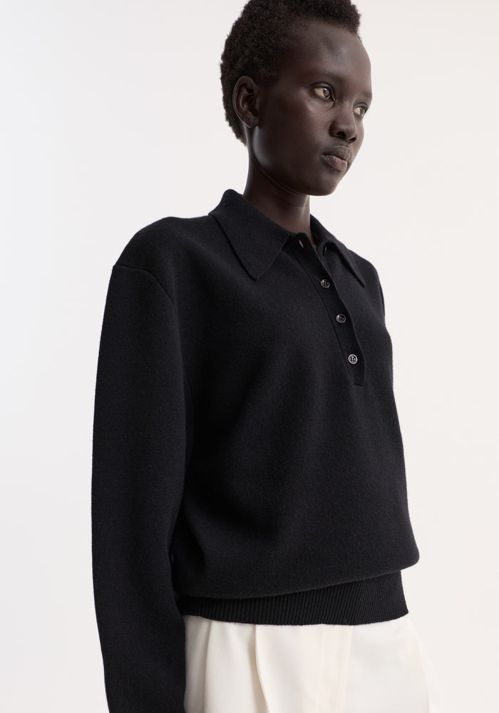 Person in a Compact knitted polo | black with a 3-button front, rounded drop shoulder, and structured collar, paired with loose white trousers.
