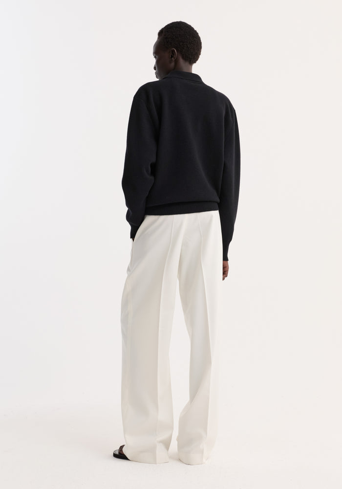 Person wearing a Compact knitted polo in black, featuring a structured collar and three-button closure, paired with wide-leg trousers, viewed from the back.