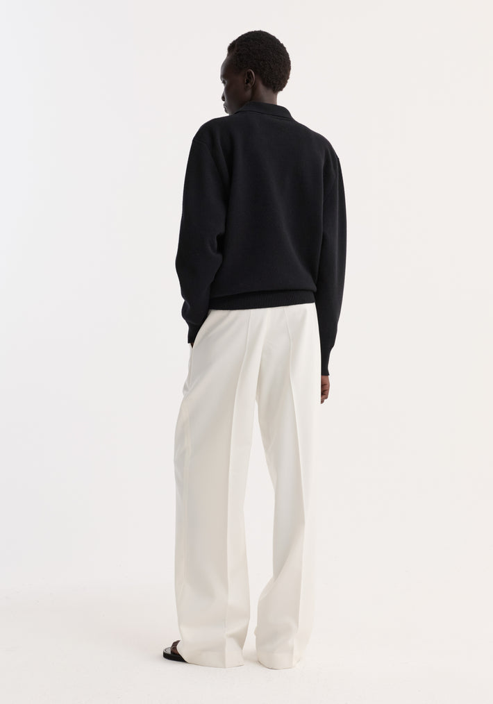 Person wearing a Compact knitted polo in black, featuring a structured collar and three-button closure, paired with wide-leg trousers, viewed from the back.