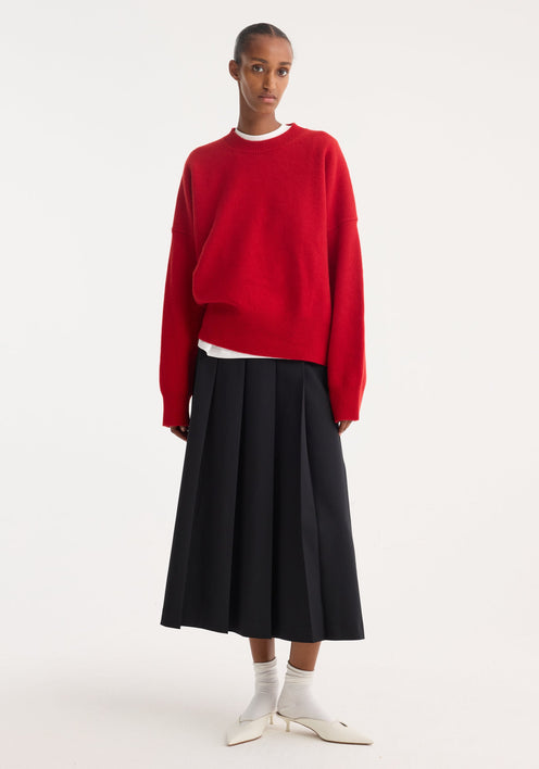 Person wearing a Boiled wool crewneck | rosso, featuring a relaxed fit and drop shoulders, paired with a black pleated midi skirt and white kitten-heel mules.