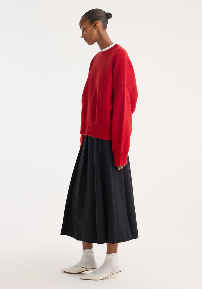 Woman in profile wearing a Boiled wool crewneck | rosso, oversized with a relaxed fit, paired with a high-waisted black pleated skirt and white mules.