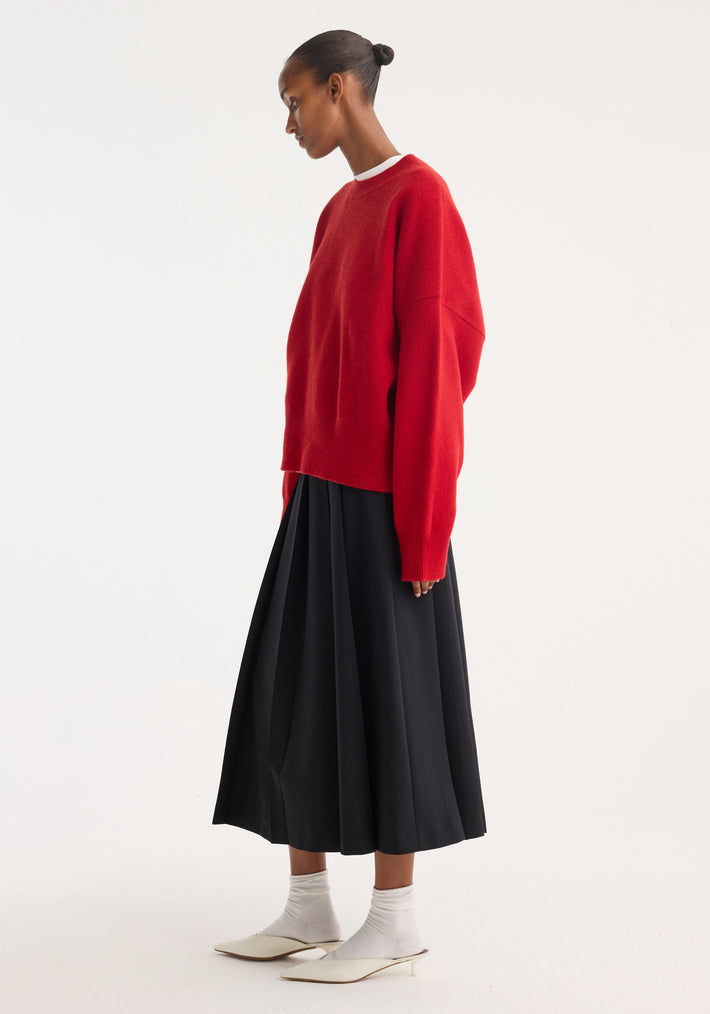 Woman in profile wearing a Boiled wool crewneck | rosso, oversized with a relaxed fit, paired with a high-waisted black pleated skirt and white mules.