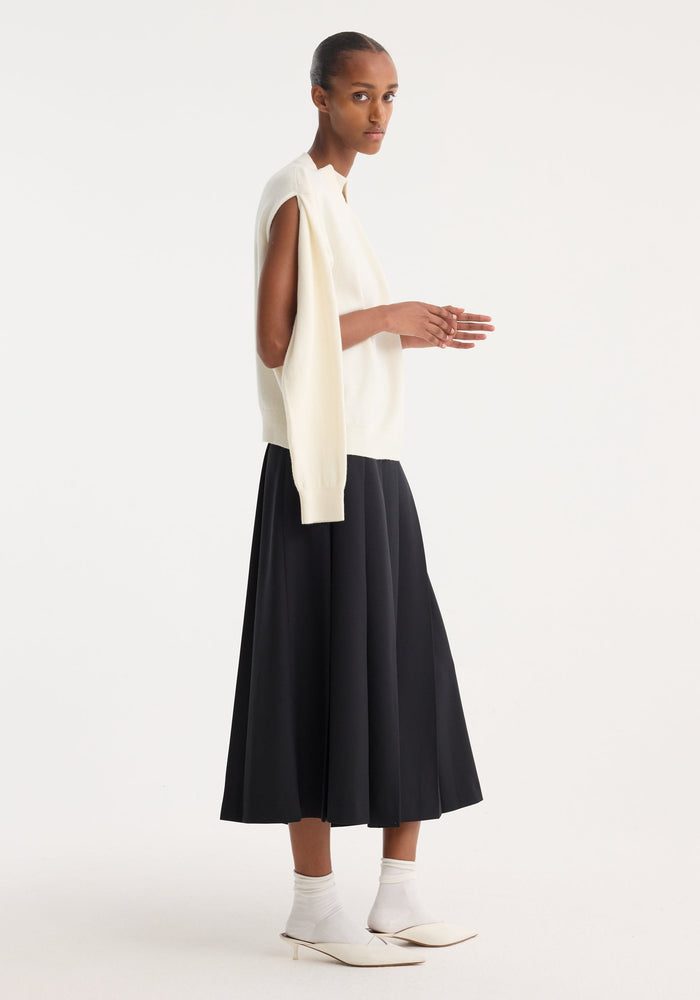 Model in profile wearing a Crewneck with open neck in off-white, featuring a deconstructed open design, loose sleeve down the side, styled with a black pleated skirt.