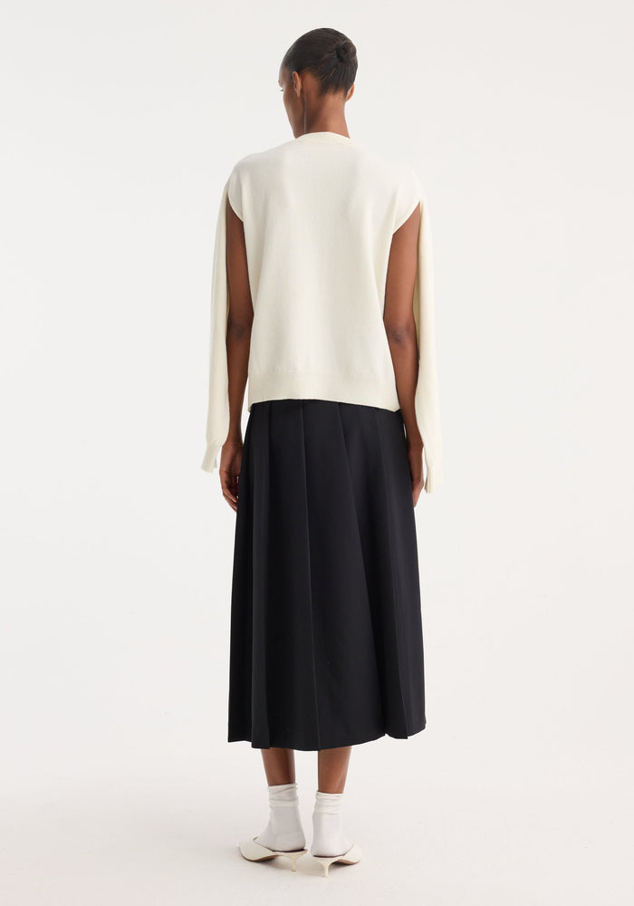 Model wearing Crewneck with open neck in off white, featuring loose, draping sleeves. Styled with a black pleated skirt, white socks, and ivory slingback shoes.