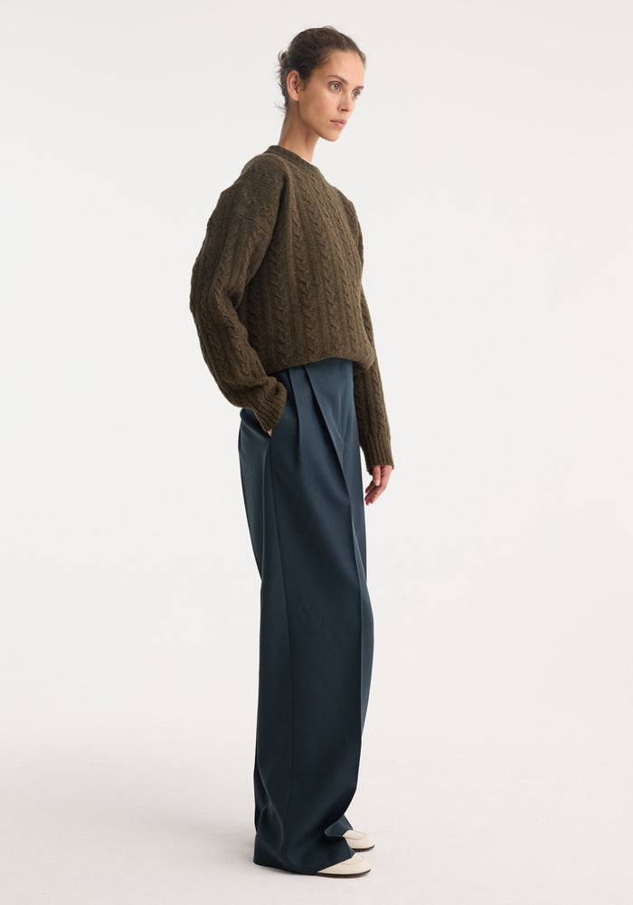 Model in profile wearing mid-waist tailored trousers | storm, featuring a tailored waistband, double pleats, and wide legs, paired with a cable-knit sweater.