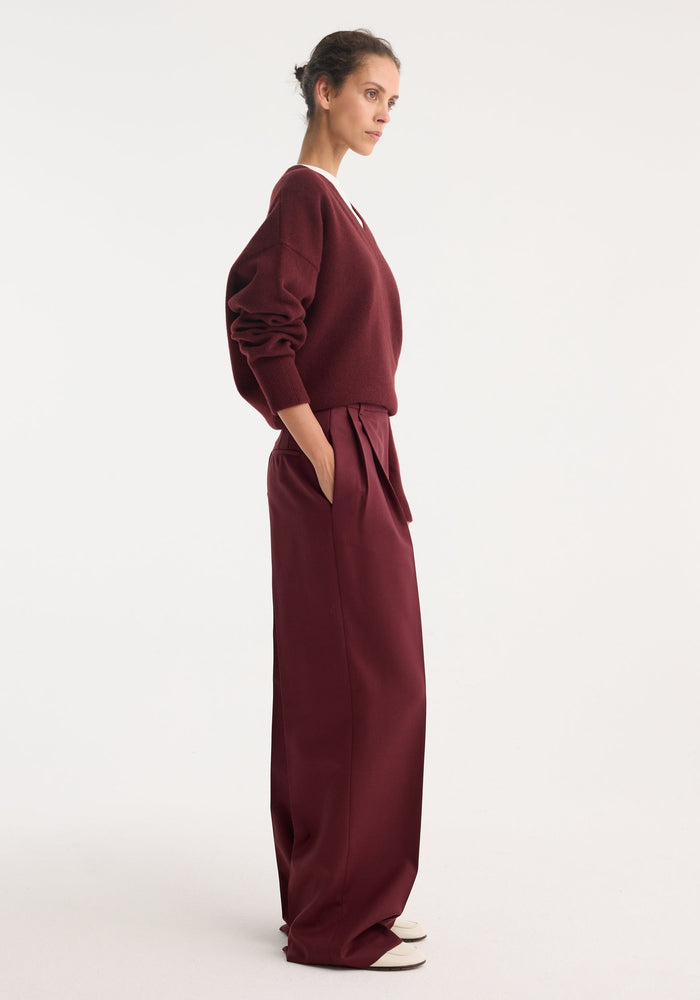 Woman in profile wearing burgundy mid-waist tailored trousers with double pleats and wide legs, styled with a V-neck sweater, hand in pocket.