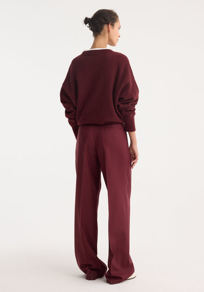 Person wearing Mid-waist tailored trousers in burgundy, featuring a wide-leg design with double pleats, seen from behind with matching sweater and white shoes visible.