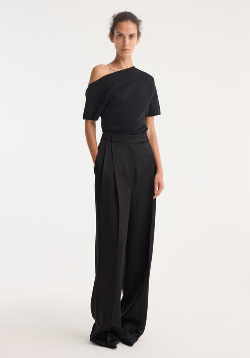 Model wearing high-waist satin trousers in black, featuring front and back pleats, welt pockets, and tailored waistband, paired with an asymmetrical knit top.