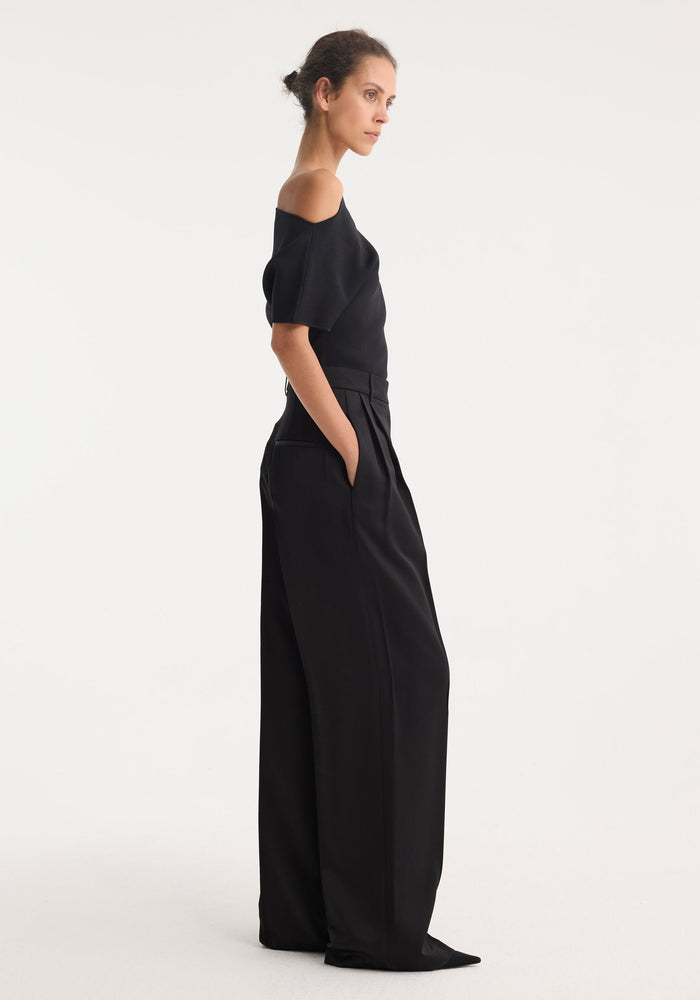 High-waist satin trousers in a side profile, featuring front and back pleats with welt pockets. Model wears them with a fitted, off-shoulder top.
