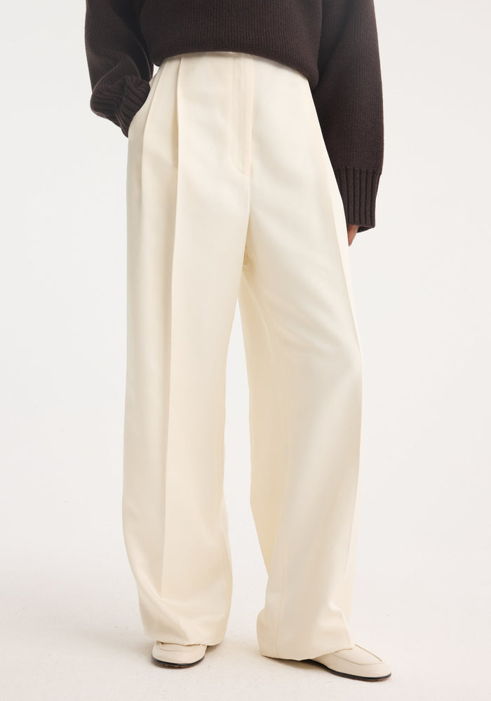 High-waist satin trousers in cream with front pleats, welt back pockets, and tailored waistband, paired with matching slip-on shoes, showcased against a plain background.