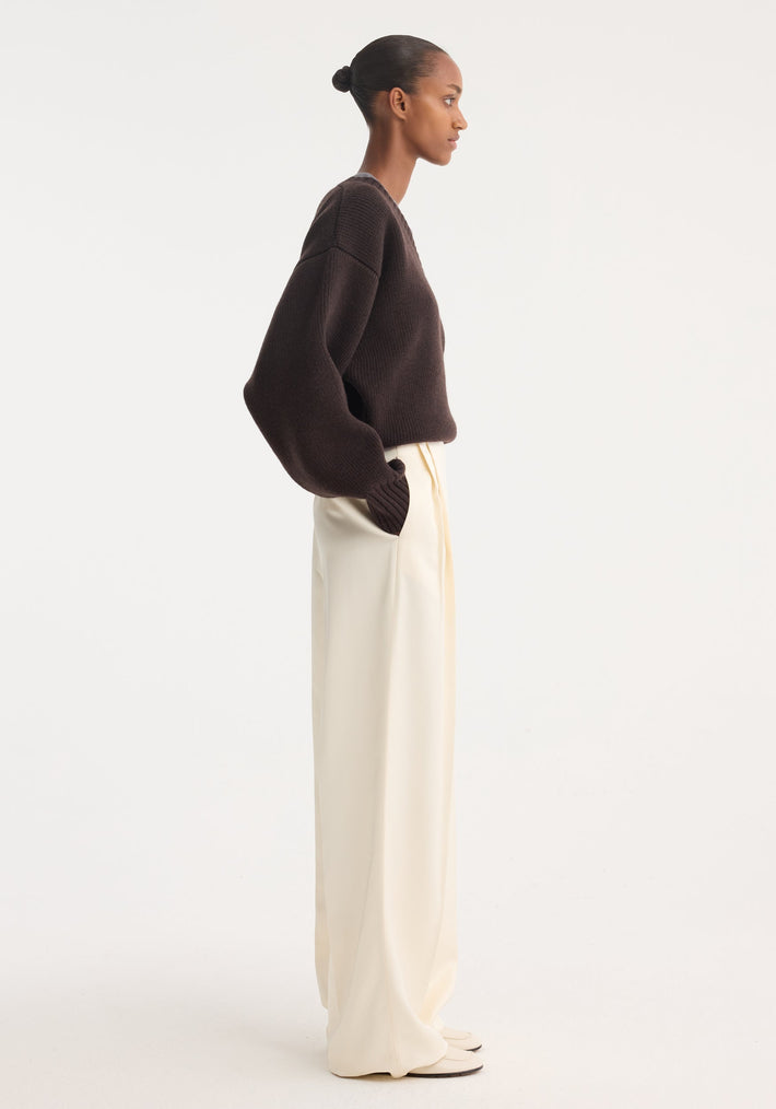 High-waist satin trousers in cream, featuring front and back pleats and welt pockets, worn by a model with hands in pockets.
