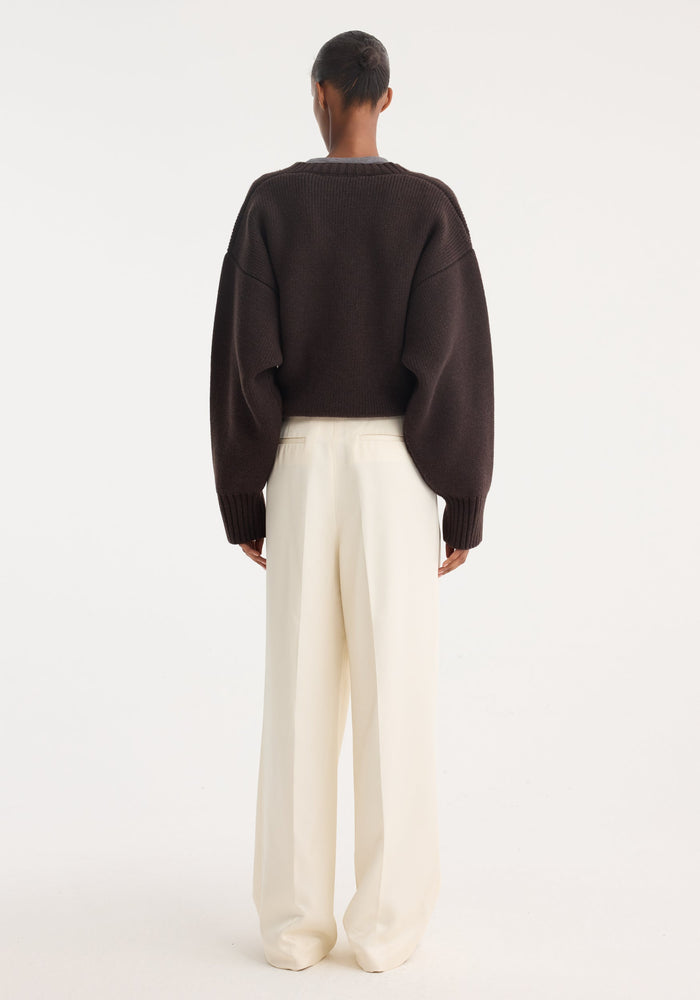 Person wearing high-waist satin trousers in cream, featuring front and back pleats, tailored waistband, and welt back pockets, paired with a chunky-knit sweater.