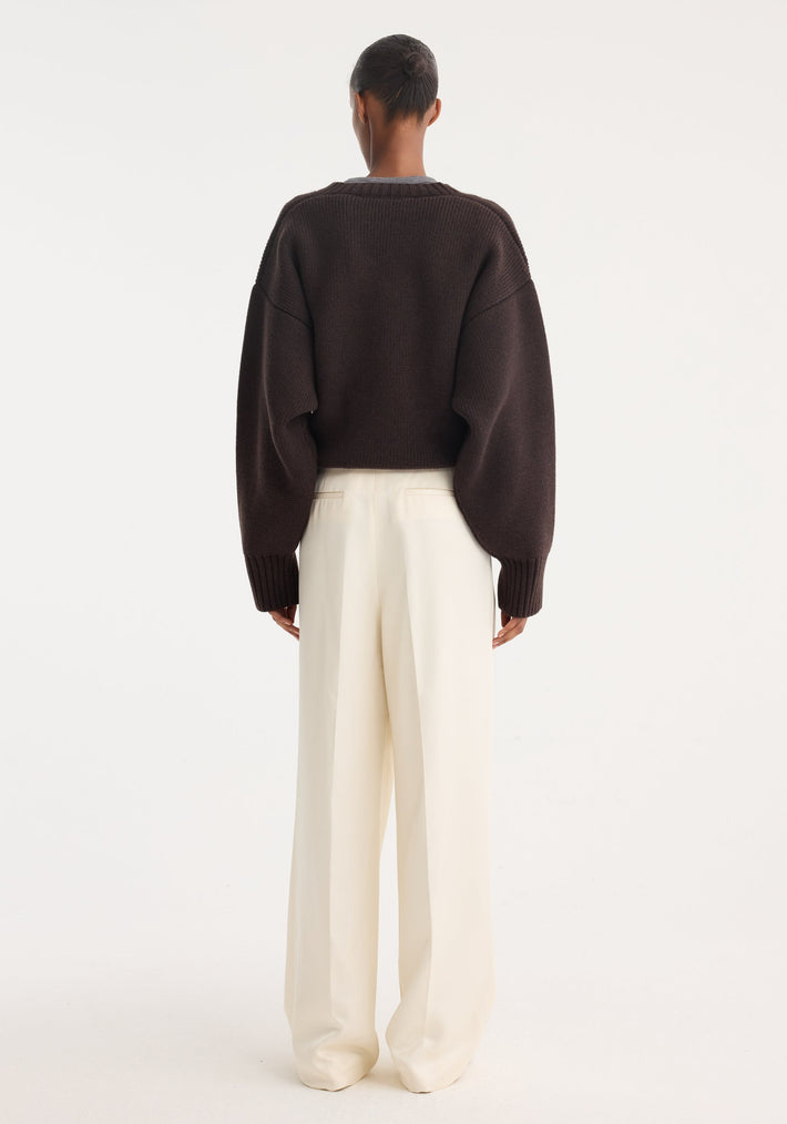 Person wearing high-waist satin trousers in cream, featuring front and back pleats, tailored waistband, and welt back pockets, paired with a chunky-knit sweater.