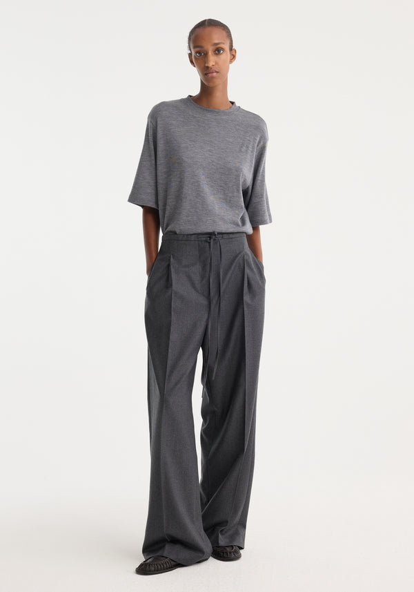 Model wearing relaxed soft wool trousers in dark grey melange, featuring wide-leg, tie-belt waist, pleats, and side pockets, showcasing elegant tailoring and comfort.