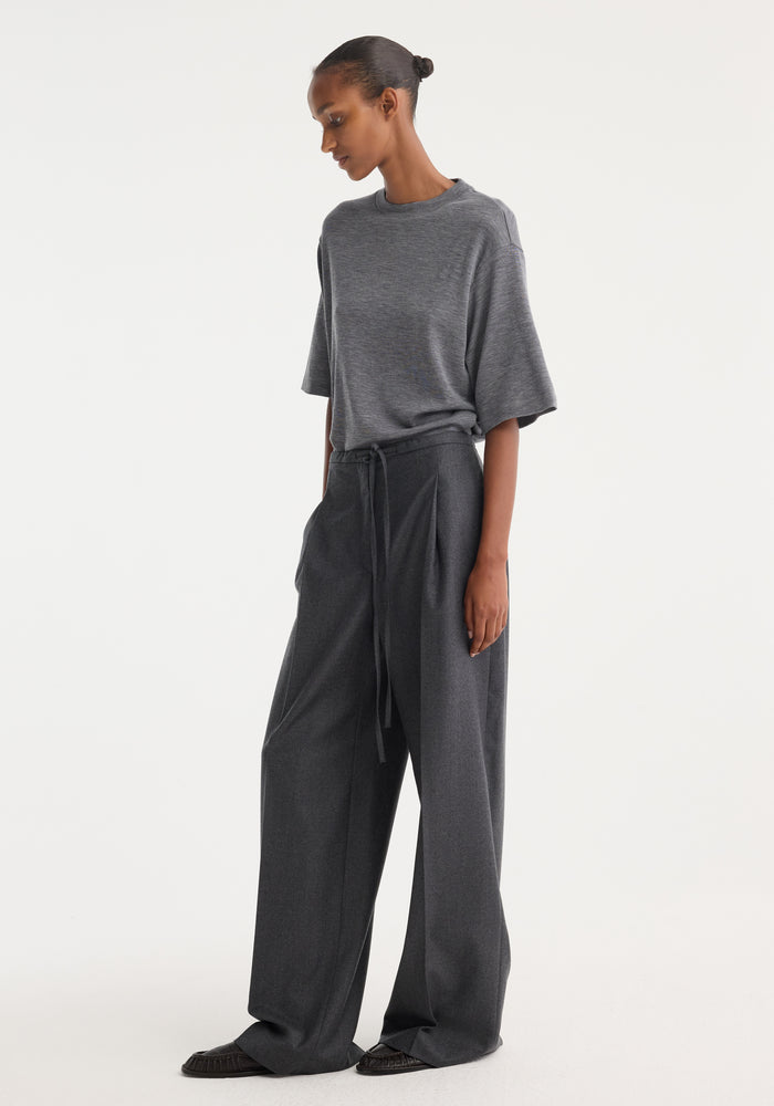 Model in Relaxed soft wool trousers with wide legs and drawstring waistband, showcasing lightweight virgin wool, paired with a charcoal-gray T-shirt and black flats.