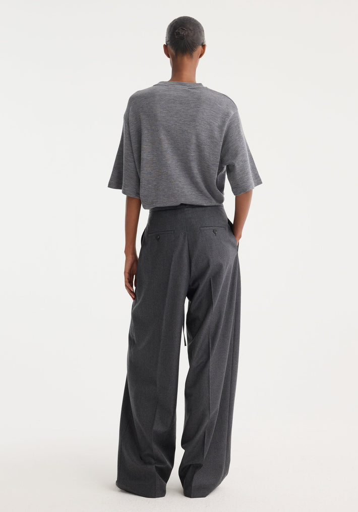Rear view of a model in Relaxed soft wool trousers | dark grey melange, showcasing a wide-leg fit with a drawstring waistband and tucked grey T-shirt.