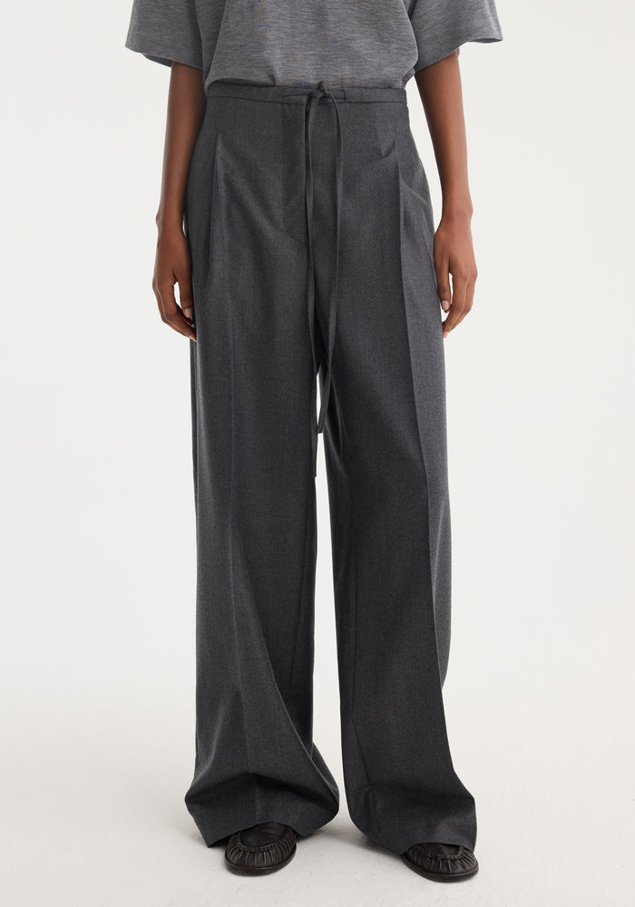 Model wearing Relaxed soft wool trousers in dark grey melange with a wide leg and drawstring waist, paired with a tucked-in T-shirt and slip-on flats.