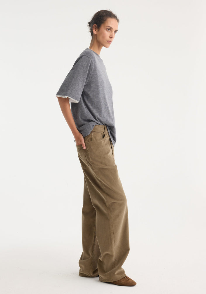 Relaxed corduroy trousers in dark sage, high-waisted with wide legs, paired with a heather-gray T-shirt. Styled with brown slip-on shoes.