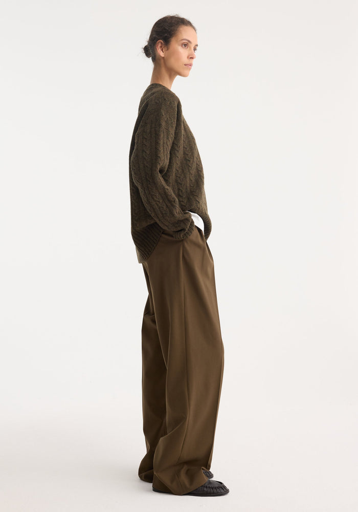 Model in profile wearing an oversized Cable knit jumper | forest melange with dropped shoulders, round neckline, paired with wide-leg trousers and black slip-on shoes.