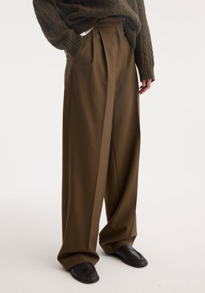 Person wearing Signature tailored trousers in forest hue, featuring wide legs and front pleats, paired with moccasin-style shoes and chunky sweater, showcasing a relaxed silhouette.