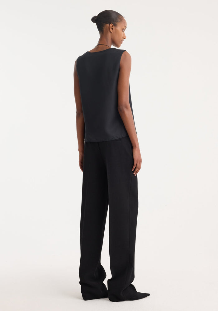Model wearing high-waisted, straight leg trousers in black, featuring sharp front pleat and tailored waistband. Made from virgin wool blend, showcasing clean side pockets.