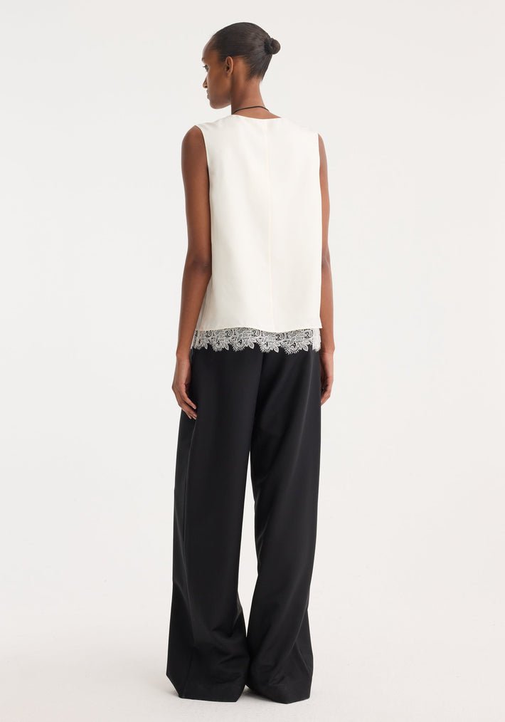 Woman in a lace-trimmed silk top | cream, with a scoop neck, sleeveless design, paired with wide-leg trousers, showcasing the blouse’s intricate lace hem.