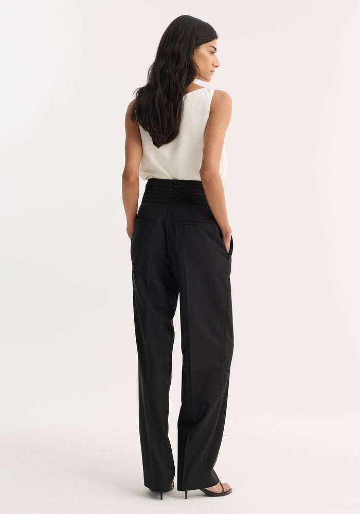 Woman in Satin Cummerbund Trousers | Black, featuring a structured waistband with shell buttons, wide legs, and slanted pockets, standing with hands in pockets.