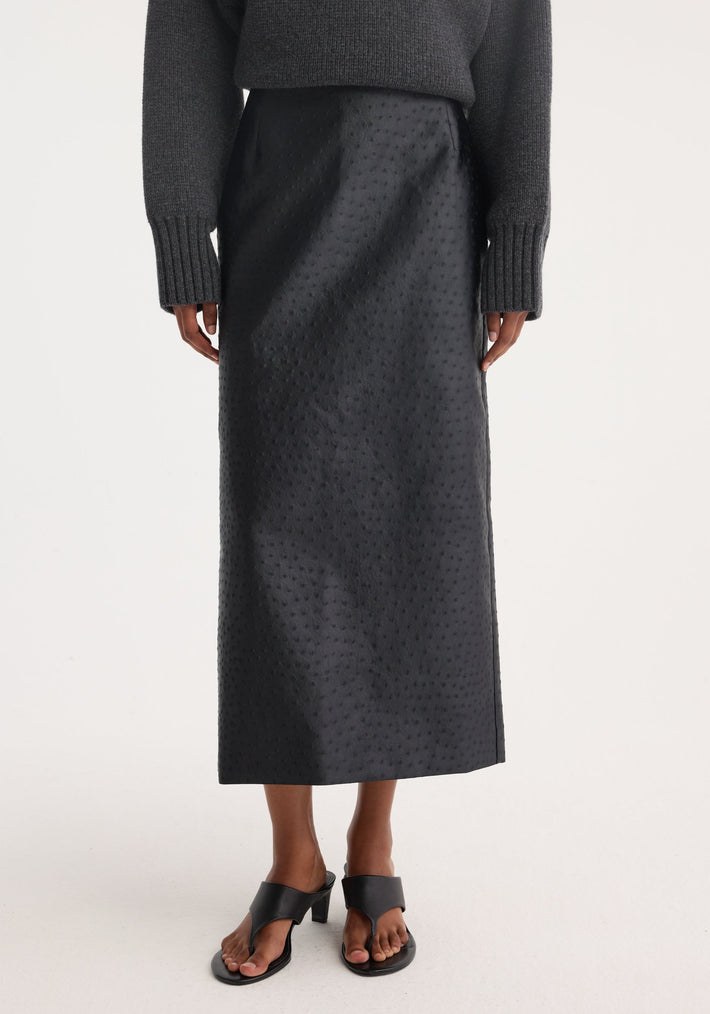 Person wearing the Ostrich satin column skirt in black, featuring an embossed ostrich-skin texture, paired with black heeled slide sandals and a charcoal knit sweater.