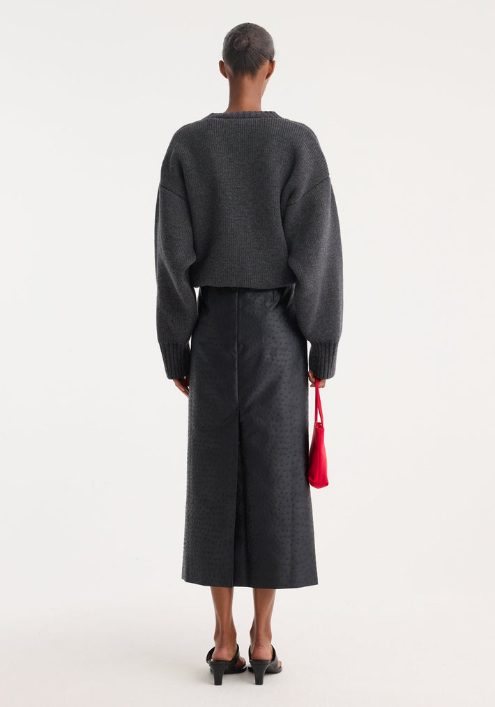 Back view of a model wearing the Ostrich satin column skirt with embossed texture, paired with a chunky knit pullover and holding a red handbag.