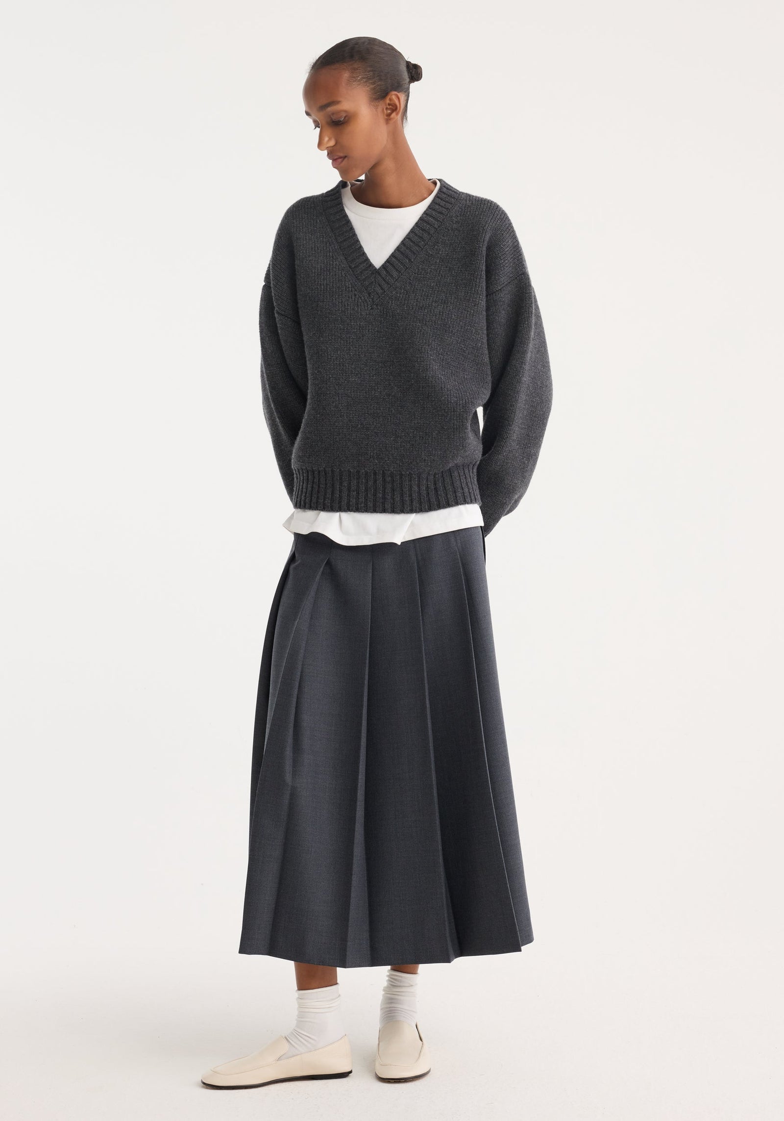 Model in a dark grey melange pleated wrap skirt with a tailored waistband and wide pleats, paired with a charcoal V-neck sweater and cream loafers.