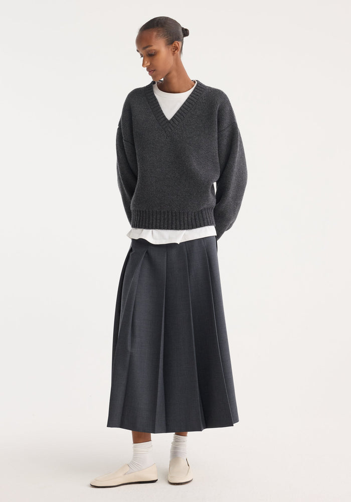 Model in a dark grey melange pleated wrap skirt with a tailored waistband and wide pleats, paired with a charcoal V-neck sweater and cream loafers.