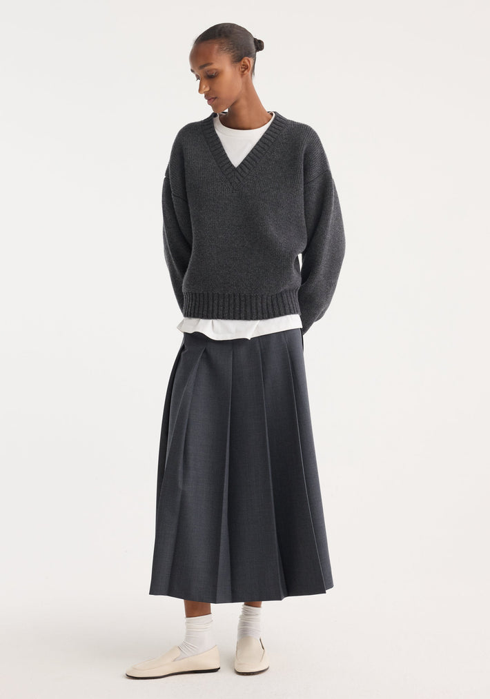 Model in a dark grey melange pleated wrap skirt with a tailored waistband and wide pleats, paired with a charcoal V-neck sweater and cream loafers.