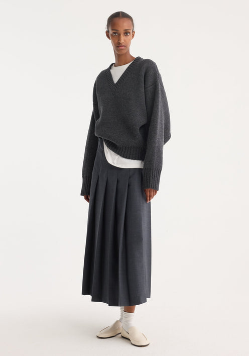 Model in a dark grey melange pleated wrap skirt with a tailored waistband and wide pleats, paired with a charcoal V-neck sweater and cream loafers.