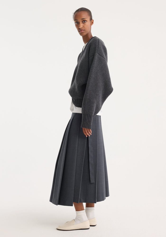 Model wearing a Pleated wrap skirt | dark grey melange, showcasing its tailored waistband, fitted hip, and wide pleats, styled with a knit sweater and loafers.