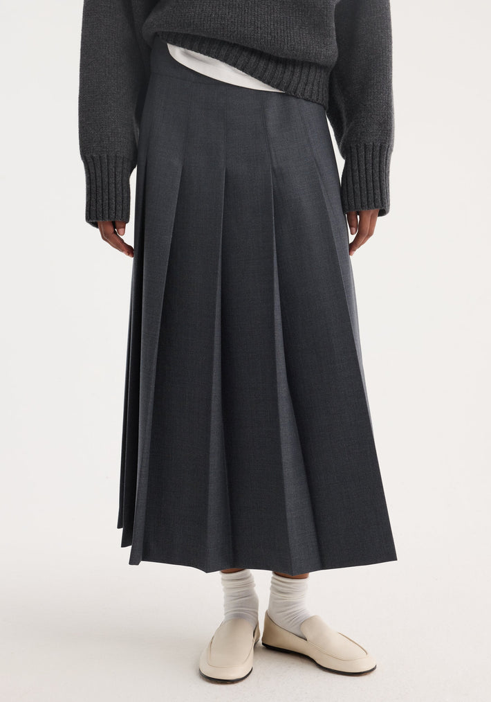 Pleated wrap skirt in dark grey melange with a tailored waistband and voluminous pleats, paired with white socks and cream loafers against a neutral background.