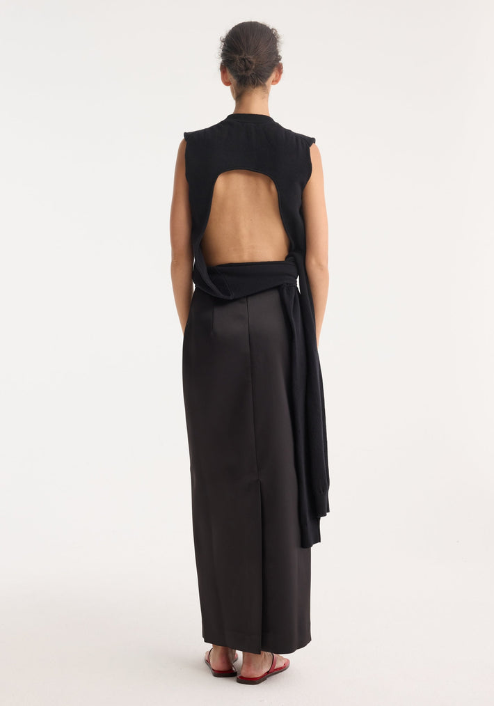 Woman in Open-back sash wrap top with sculptural sleeves tied at waist, long black skirt, and red sandals, showcasing a minimalist, modern fashion look.