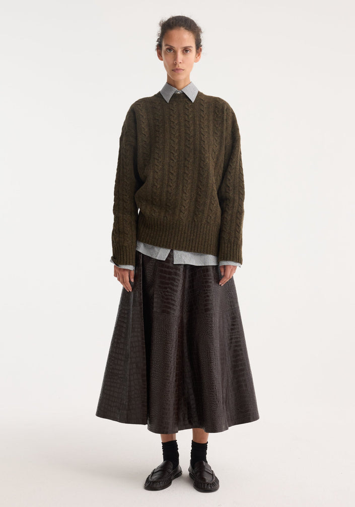 Model wearing the A-line croco skirt in mahogany, featuring a glossy, embossed finish, paired with a green knit sweater and loafers.
