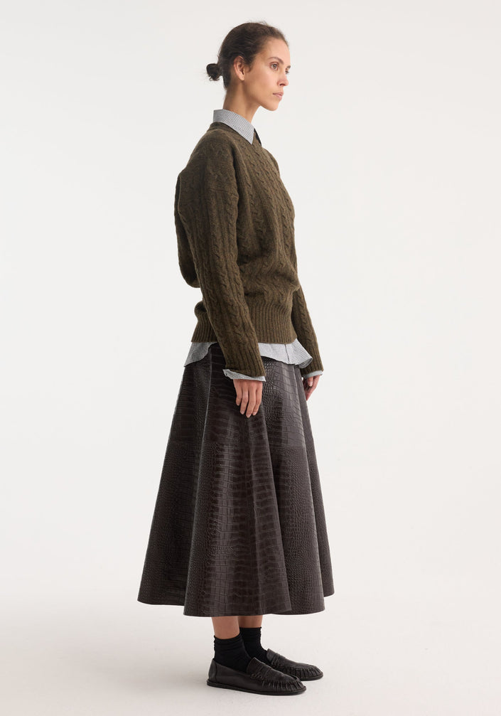 Model wearing the A-line croco skirt in mahogany, featuring a glossy, embossed finish, paired with a green knit sweater and loafers.