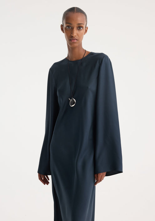 Model in Signature silk dress | storm, featuring a full-length, navy column silhouette with wide sleeves, accessorized with a black cord necklace and sandals.