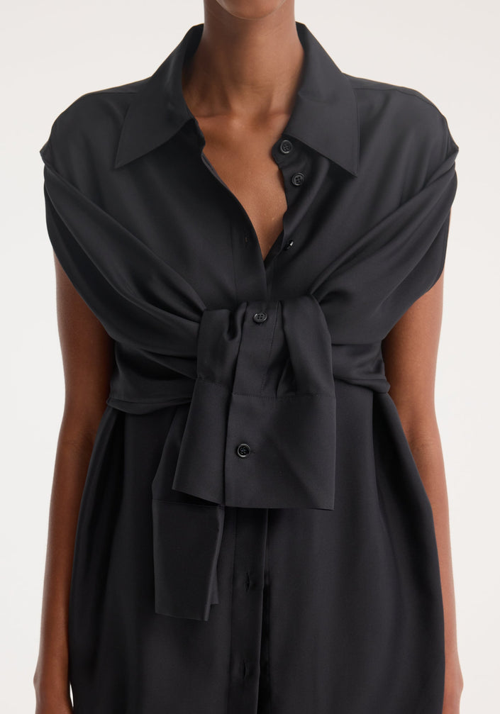 Model wearing a Silk gilet-dress in black, featuring a deconstructed design with open armholes, buttoned wrap belt, and fluid silhouette. Paired with black heeled sandals.