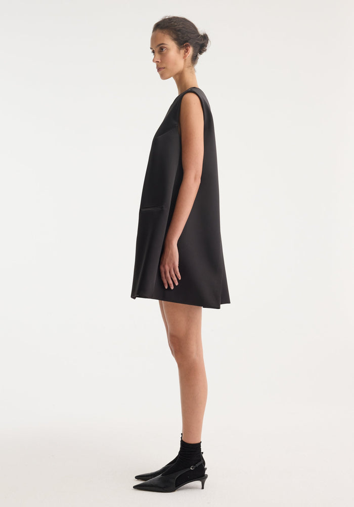 Woman in Sculptural cape dress | black, side profile. Sleeveless, A-line with hip welt pocket, pointed-toe kitten heels, neat bun. Hidden tie for tailored fit.