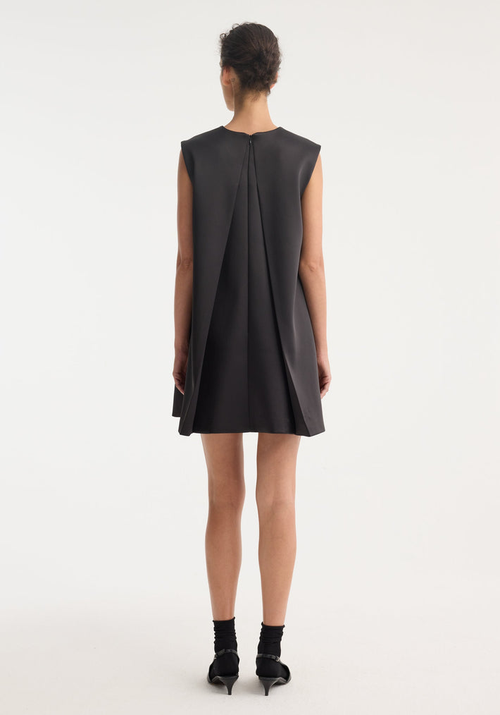 Woman in Sculptural cape dress | black, showcasing a sleeveless A-line mini with dramatic back box pleats, mid-calf socks, and low-heeled Mary-Jane shoes.